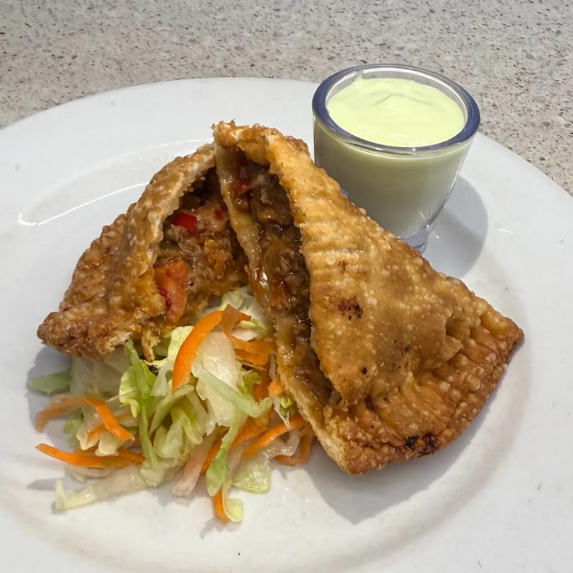 Two golden-brown empanadas, one cut open to show a savory filling, served with a side salad and green sauce.