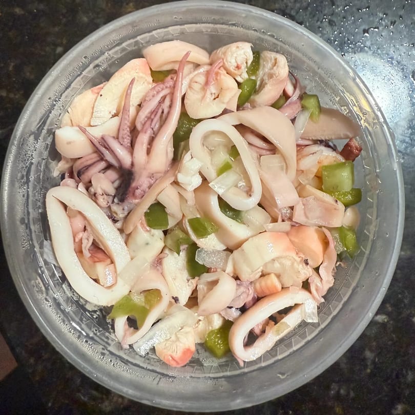 A clear plastic container filled with Angiolina's Restaurant's Latin-American seafood salad, featuring squid, shrimp, and green peppers.