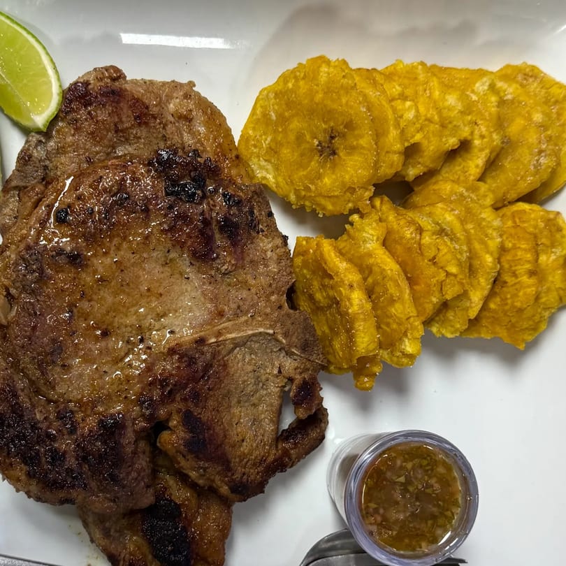 Grilled steak with a lime wedge, crispy plantain chips, and a side of sauce on a white plate.